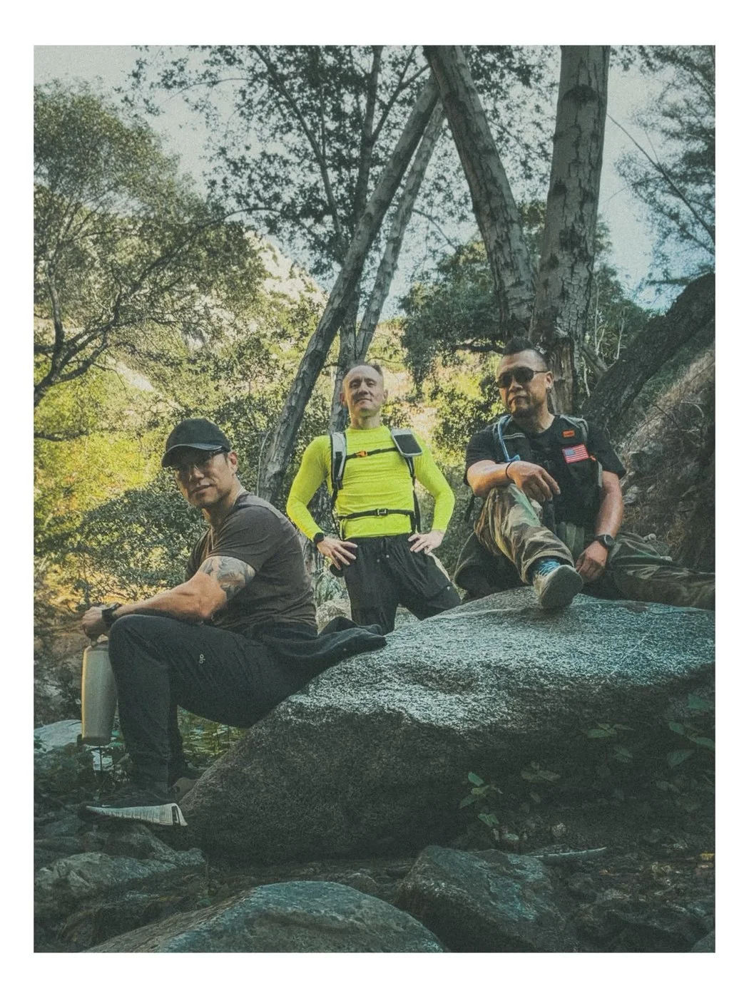 Three hikers taking a break on a boulder beneath trees.