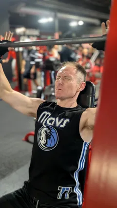 A man wearing a black basketball jersey exercising on a resistance machine.